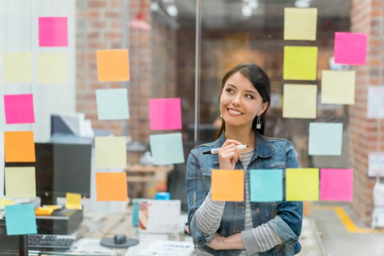 A smiling woman stands and reviews a wall of colorful post-it notes, examining her plan for social media for nonprofits.