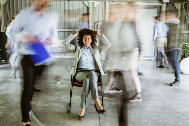 Troubleshooting WordPress websites can be frustrating! A woman sits on a chair, pulling at her hair in frustration while surrounded by blurred, fast-paced images of colleagues.