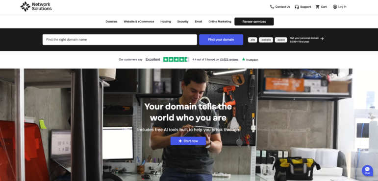 A person sits in a workspace using a smartphone, with a computer and tools around. The Network Solutions website is shown, featuring nonprofit website builders, a domain search bar, and a Start now button for domain registration services.
