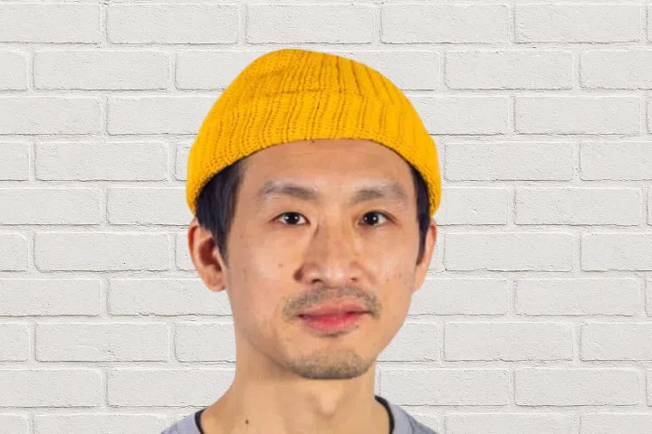 A man wearing a yellow knit beanie and a light gray shirt stands in front of a white brick wall.