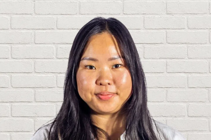A woman with long dark hair and wearing a white top is standing in front of a white brick wall, looking directly at the camera with a neutral expression.