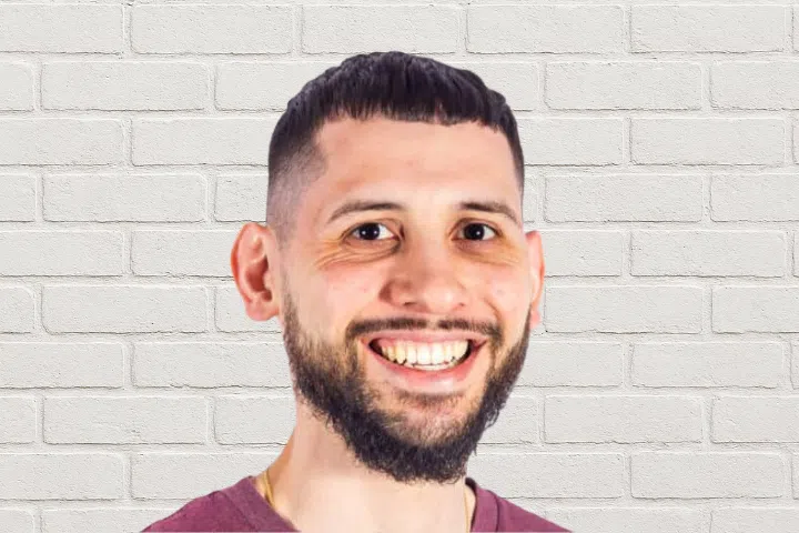 A man with short dark hair, a trimmed beard, and a maroon shirt is smiling at the camera. He is standing in front of a white brick wall background.