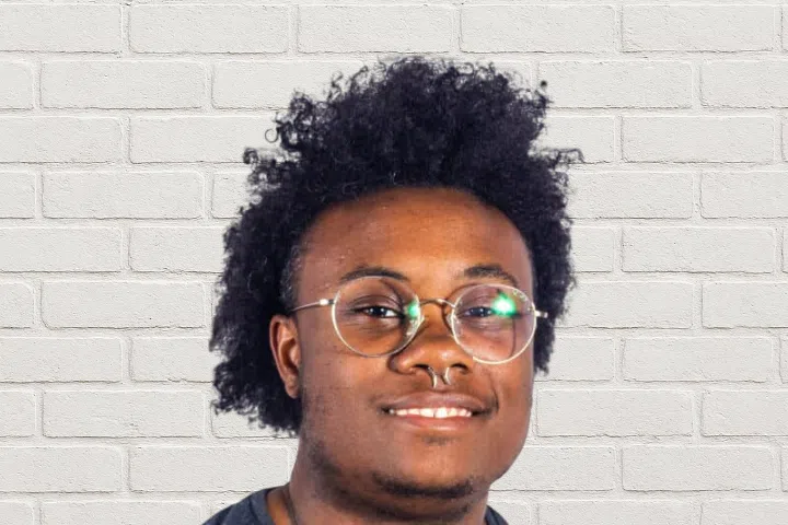 A person with short curly hair, round glasses, and a nose ring is smiling in front of a white brick wall background.