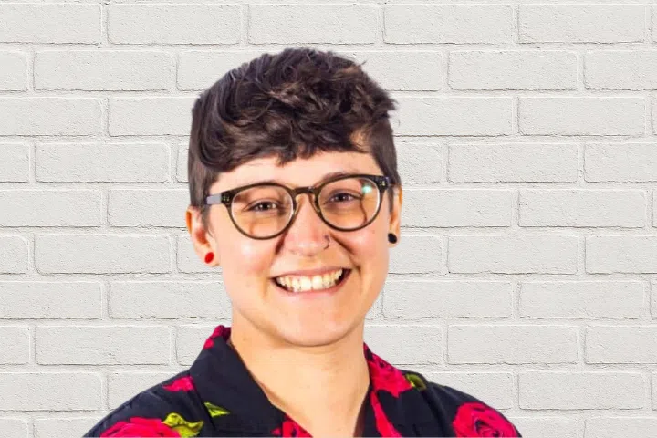 A person with short, curly brown hair and glasses smiles in front of a white brick wall. They are wearing a black shirt with a red and green floral pattern and red stud earrings.