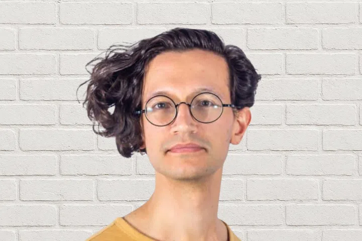 A person with wavy, medium-length hair and round glasses stands in front of a white brick wall, wearing a light brown shirt.