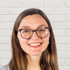A woman with long brown hair and glasses is smiling in front of a white brick wall background. She is wearing a gray top.