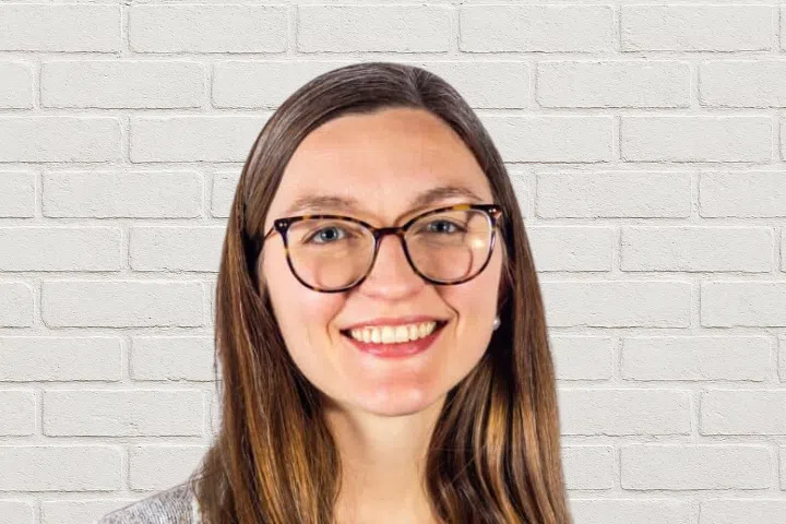 A woman with long brown hair and glasses is smiling in front of a white brick wall background. She is wearing a gray top.