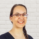 A person with long brown hair in a braid, wearing glasses with red frames and a black top, smiles in front of a white brick wall.