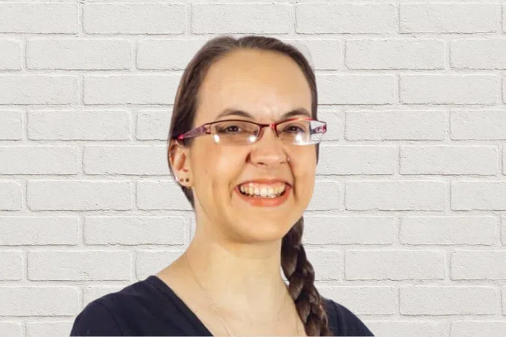 A person with long brown hair in a braid, wearing glasses with red frames and a black top, smiles in front of a white brick wall.