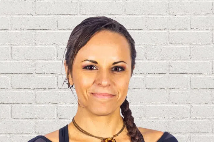 A person with dark hair in a braid, wearing a black top and a necklace, smiles slightly while standing in front of a white brick wall.