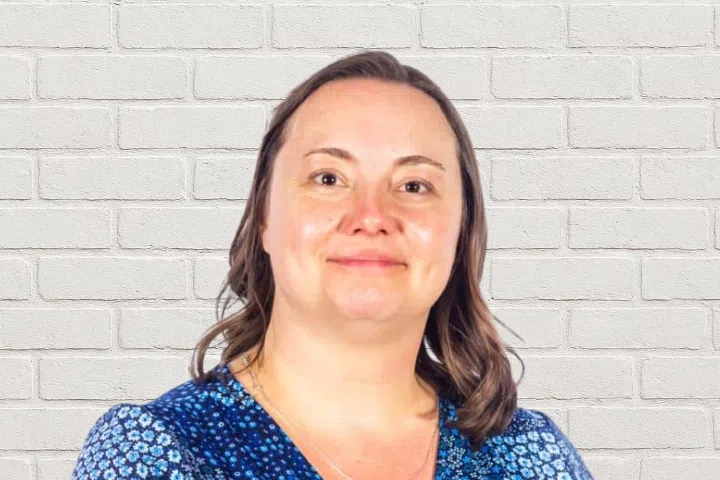 A woman with shoulder-length brown hair, wearing a blue patterned top, is standing in front of a white brick wall and looking at the camera with a neutral expression.