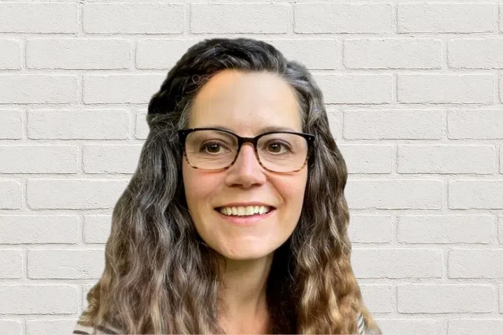 A woman with long, wavy brown and gray hair, wearing glasses and a striped top, smiles at the camera in front of a light-colored brick wall background.