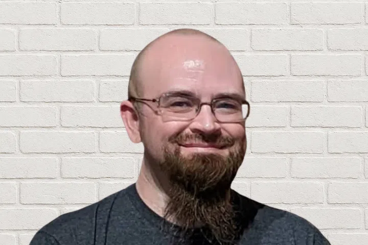 A man with a bald head, glasses, and a long brown goatee is smiling. He is wearing a dark t-shirt and standing in front of a white brick wall.