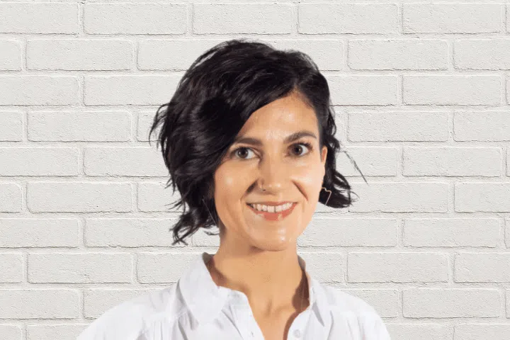 A person with short, wavy dark hair and a white collared shirt stands in front of a white brick wall, smiling at the camera.