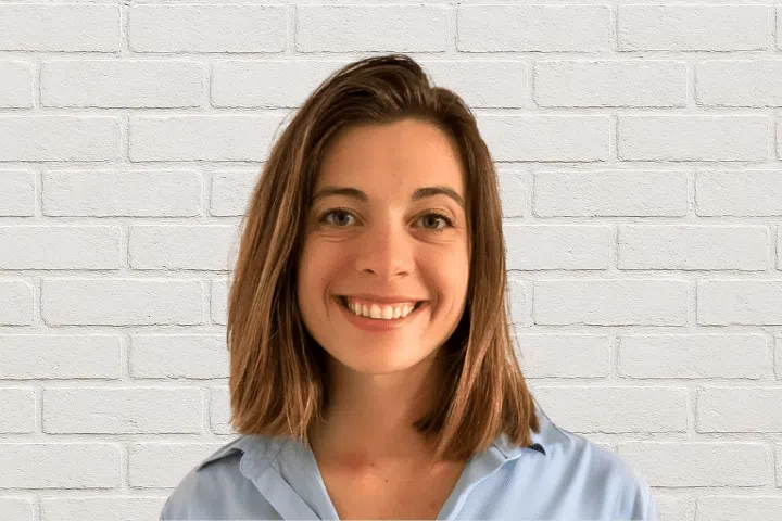 A woman with shoulder-length brown hair, wearing a light blue collared shirt, smiles at the camera while standing in front of a white brick wall.