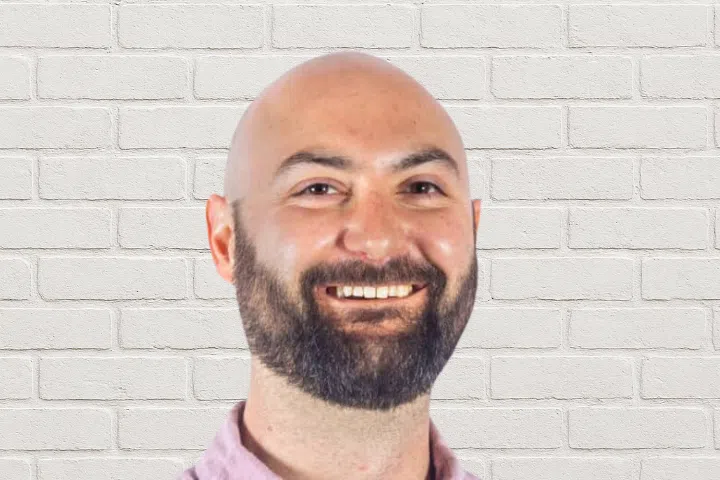 A smiling bald man with a full beard is in front of a white brick wall, wearing a light pink collared shirt.