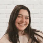 A woman with long dark hair and a light-colored sweater smiles in front of a white brick wall background.