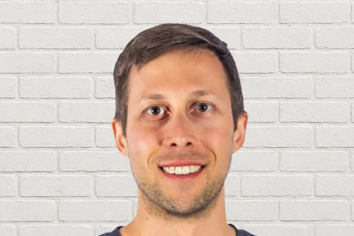A man with short brown hair and light skin is smiling while standing in front of a white brick wall.