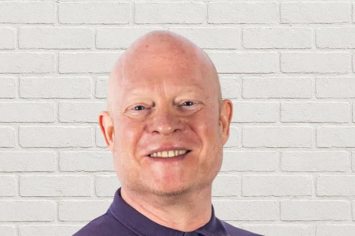 A smiling bald man wearing a dark collared shirt stands in front of a white brick wall.