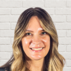 A woman with long, wavy blonde hair smiles at the camera. She is standing in front of a white brick wall background.