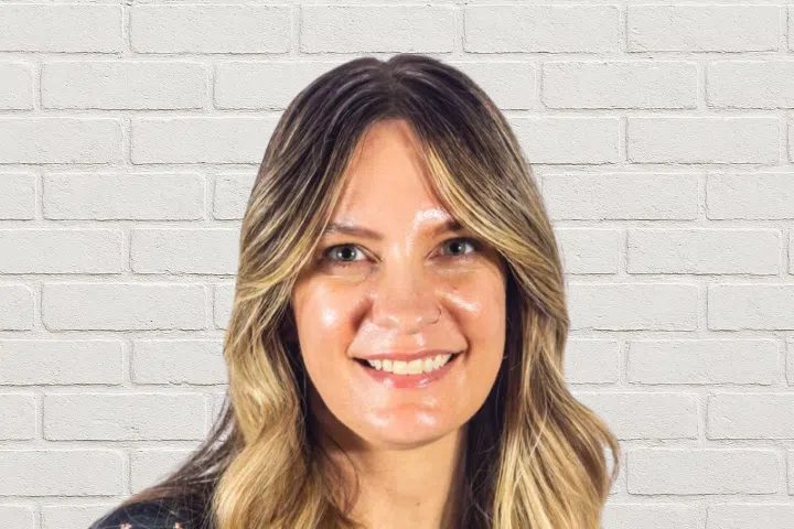 A woman with long, wavy blonde hair smiles at the camera. She is standing in front of a white brick wall background.