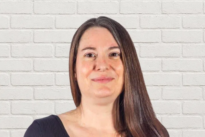 A woman with straight brown hair and a black top is smiling slightly in front of a white brick wall.