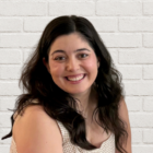 A person with long dark hair sits in front of a white brick wall, smiling at the camera and wearing a sleeveless, light-colored checkered top.